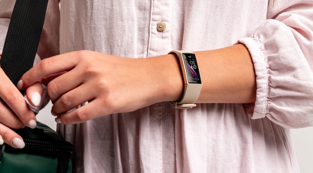 A person wearing a light, buttoned dress holds a green bag with black straps. They are wearing a smartwatch, and the background is plain.