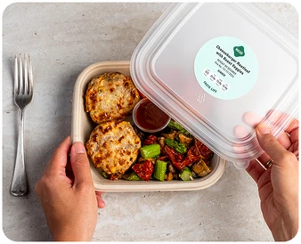 A person opens a meal container holding two cheese-topped muffins, roasted vegetables, and sauce in a cup. The label reads: Cheesy Veggie Melt with Spaghetti Squash Marinara with nutrition details. A fork lies nearby.
