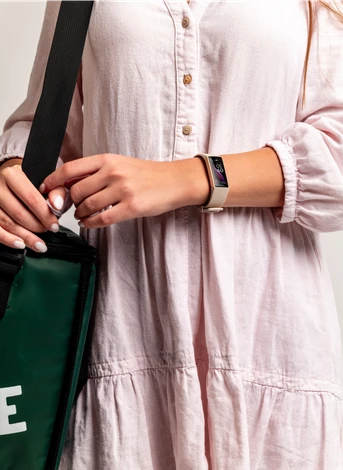 A person wearing a light, buttoned dress holds a green bag with black straps. They are wearing a smartwatch, and the background is plain.