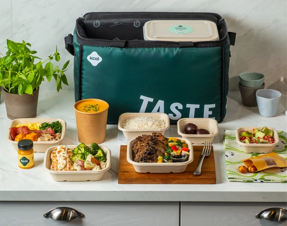 Lunch containers filled with various meals and snacks are arranged on a kitchen counter. A large insulated bag with TASTE and kcal logos is in the background, alongside potted herbs.