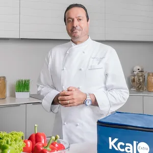 Arab man standing in a kitchen with a Kcal Meal Plans bag surrounded by healthy ingredients.