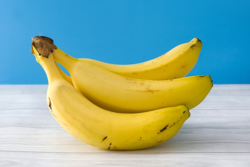 A bunch of 3 bananas sitting on a white wooden tabletop against a blue backdrop