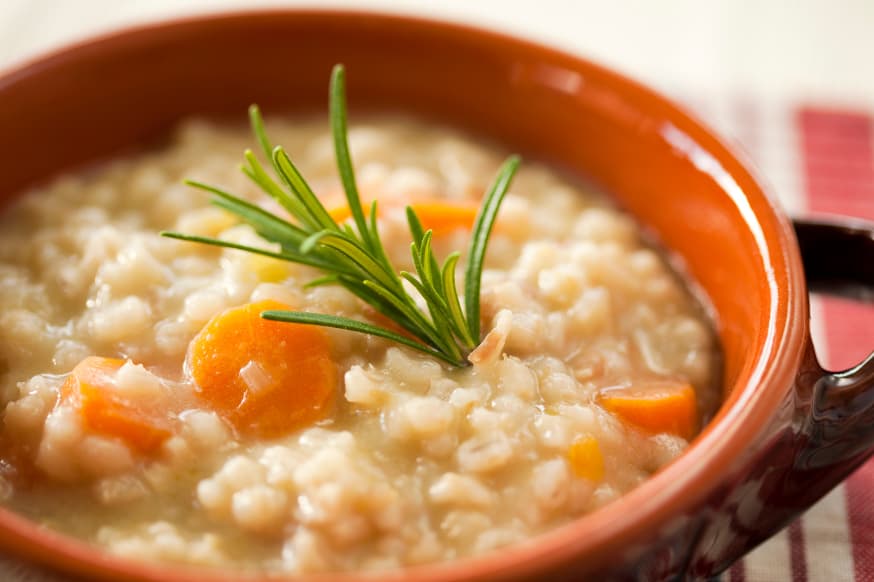 Gut-healthy vegetable soup with barley in a brown ceramic bowl on a table.