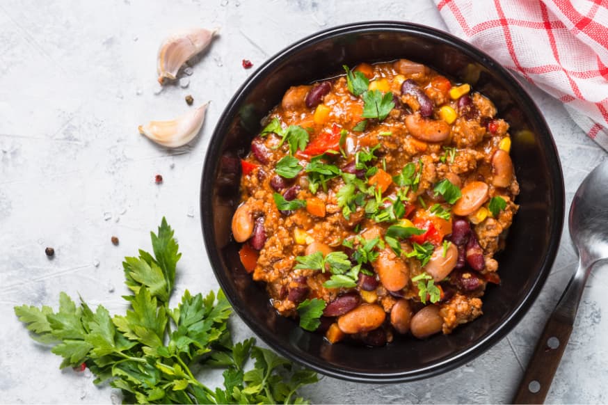 Mixed bean casserole in a black bowl on a white stone tabletop with fresh garlic cloves