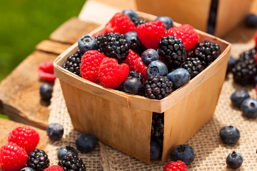 A square container of mixed berries on a wooden benchtop outside on the grass