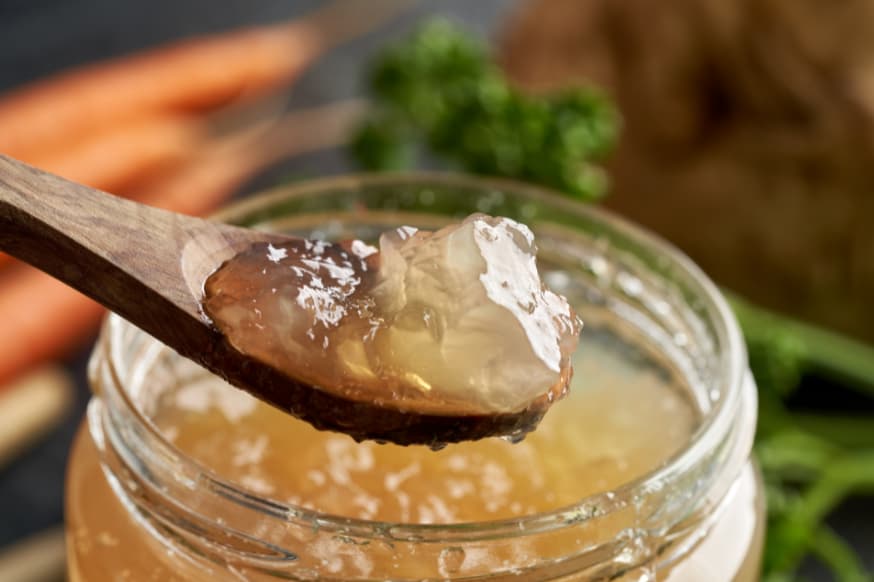 A wooden spoon filled with gelatinous bone broth scooped from a glass jar of bone broth. 