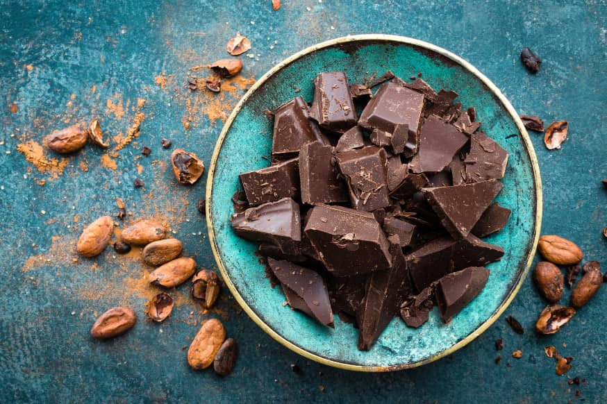 A crumbled bar of dark chocolate in a blue bowl with raw cocoa nibs on the table