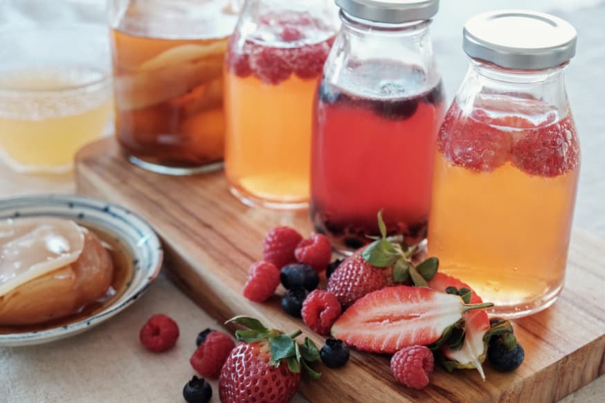 Four glass jars of a variety of kombucha teas on wooden chopping board surrounded by fresh fruits