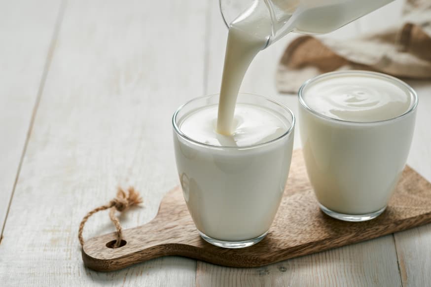 Two glasses of plain kefir being poured onto a small wooden serving tray