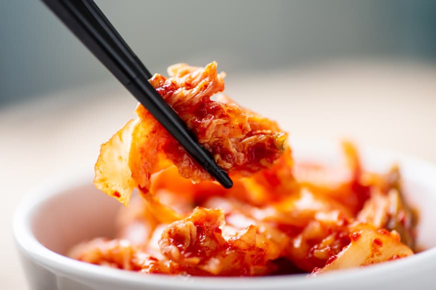 Kimchi being picked up from a small white bowl by a set of dark chopsticks