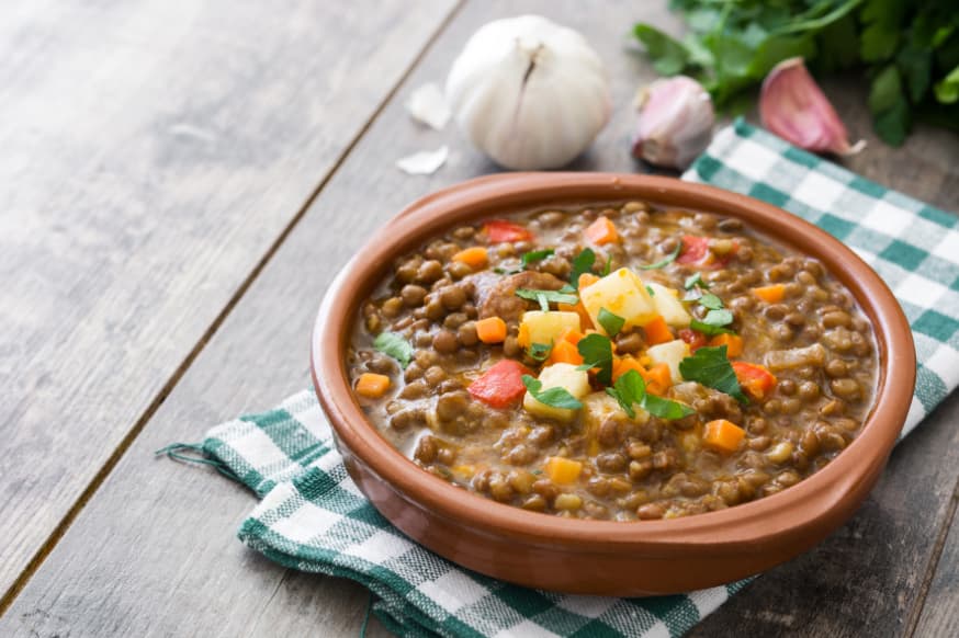 Lentil soup served in a brown ceramic bowl on a wooden table with a green and white serviette with fresh garlic next to it