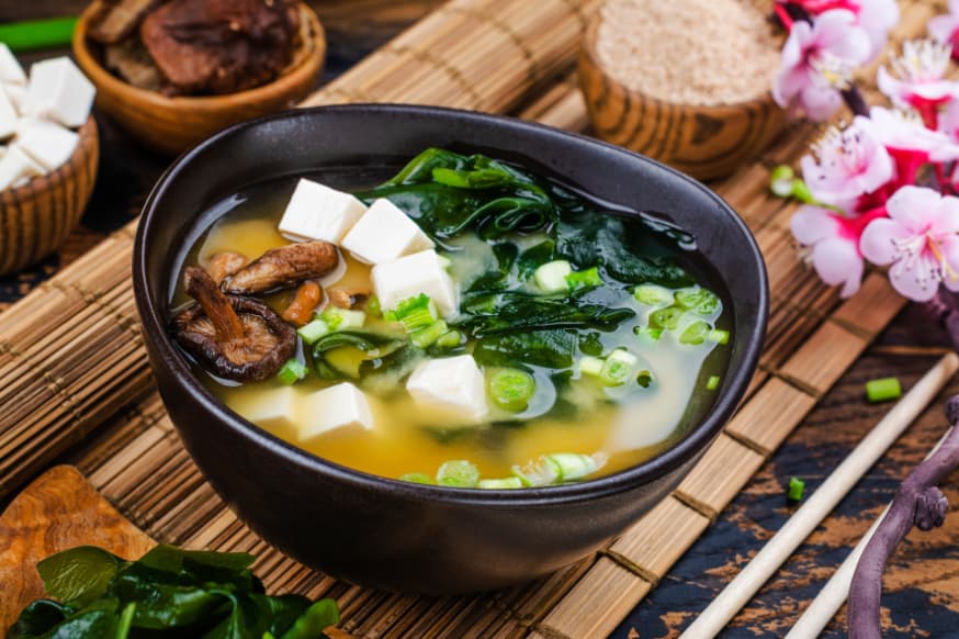 A black bowl of miso soup with seaweed, tofu cubes and mushrooms placed on a bamboo serving mat