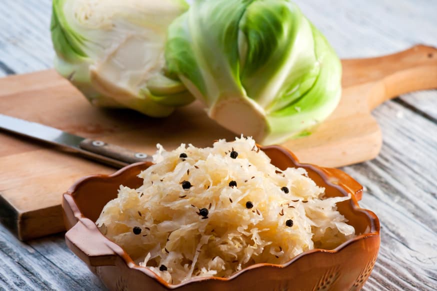 Sauerkraut in a bowl with a chopping board and raw cabbage