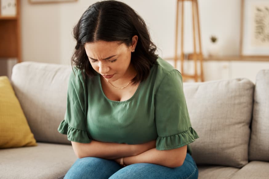 Healthy woman sitting on the couch holding her gut looking like she is in discomfort.