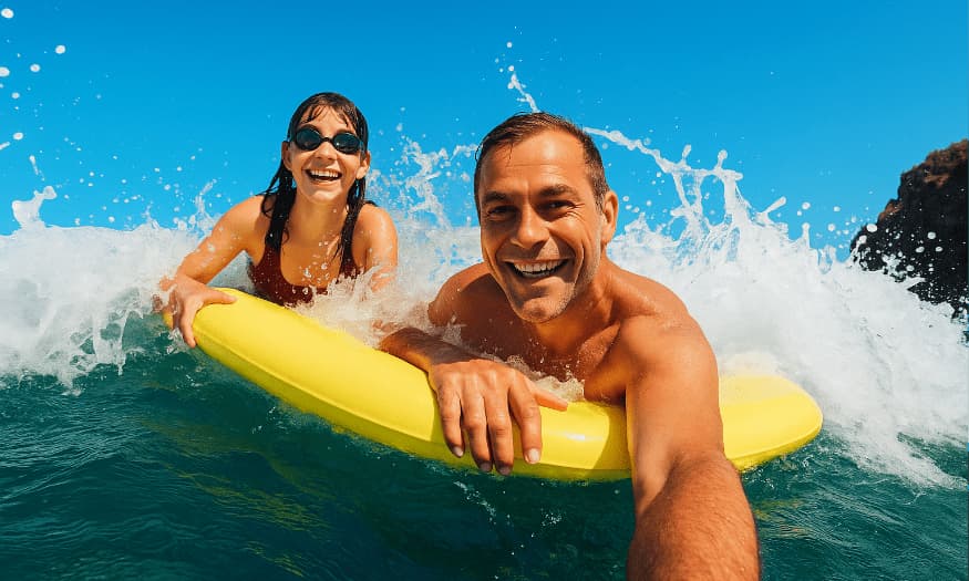 A smiling man and woman riding a yellow float board in the ocean, splashing through waves under a bright blue sky.