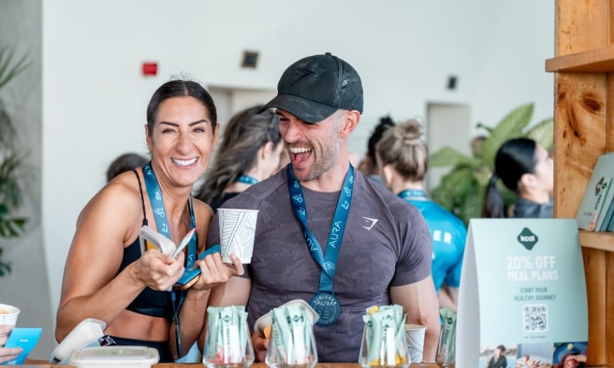 Two smiling athletes wearing medals stand at a counter, holding drinks and snacks after an event, laughing and enjoying themselves.