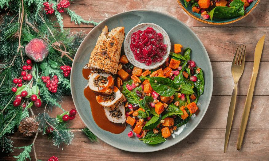 Festive plate of sliced stuffed chicken with gravy, cranberry sauce, and a spinach salad with roasted squash and pomegranate seeds on a rustic wooden table.