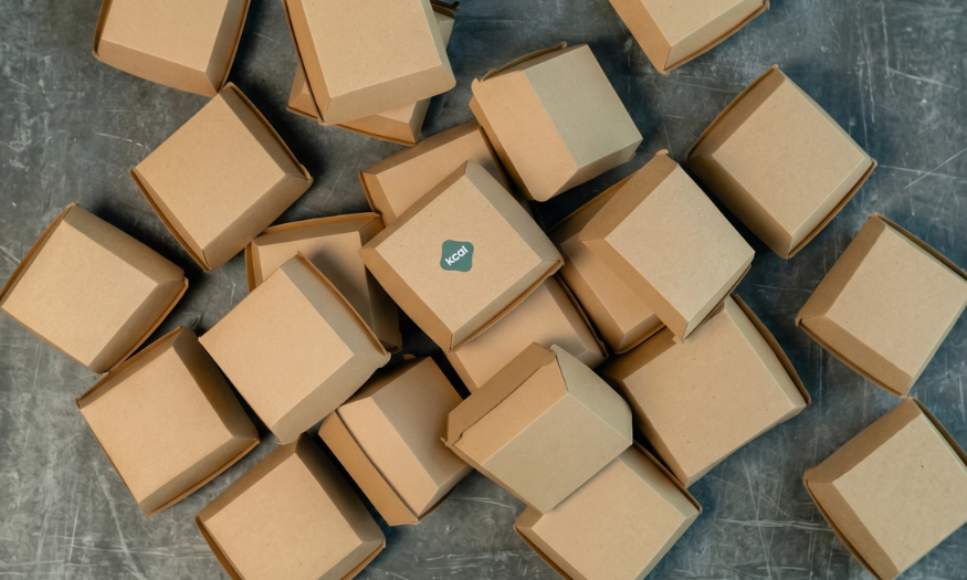 A top-down view of numerous closed, square kraft paper food containers scattered across a grey, textured industrial surface, with one central box featuring a small green "kcal" logo.